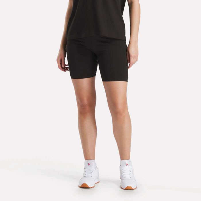 A person wears Reebok Women's Small Logo Cycle Shorts, a black T-shirt, and white sneakers against a plain white background. Only the lower half of the body is visible.
