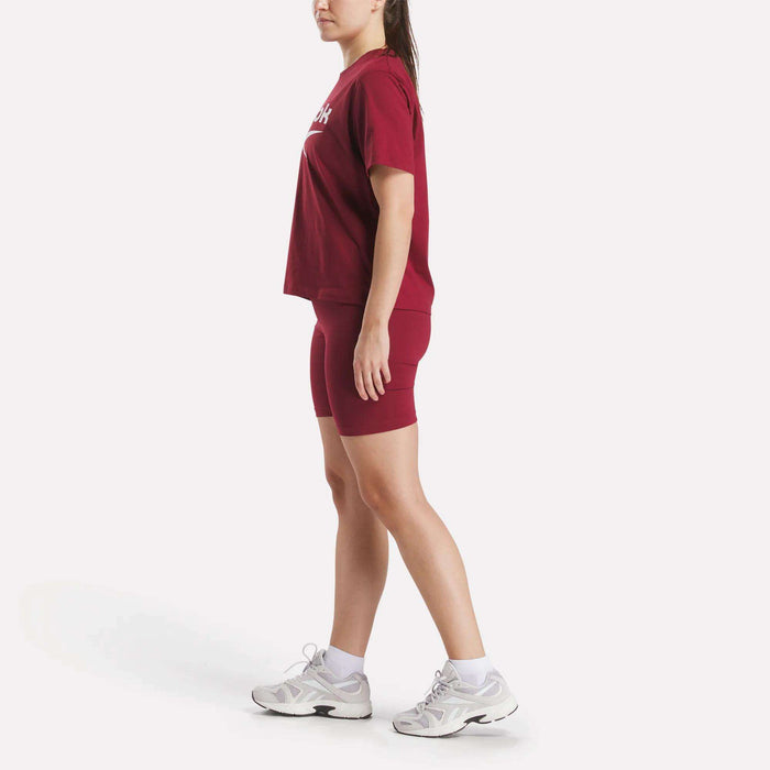 A woman stands in profile wearing Reebok Women's Small Logo Cycle Shorts with a matching red T-shirt, white sneakers, and ankle socks against a plain white background.