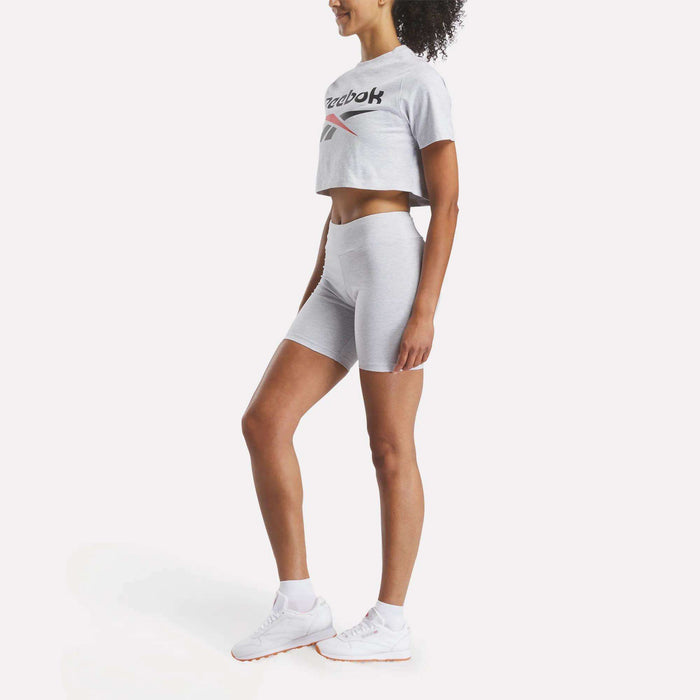 A woman stands sideways wearing light grey Reebok Women's Small Logo Cycle Shorts, a matching cropped t-shirt, and white sneakers against a plain white background.