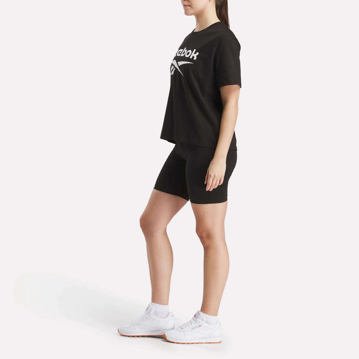 A person stands on a white background wearing a black Reebok t-shirt, Women's Reebok Small Logo Cycle Shorts with a stretchy fit, and white sneakers, facing slightly to the side with arms relaxed at their sides.