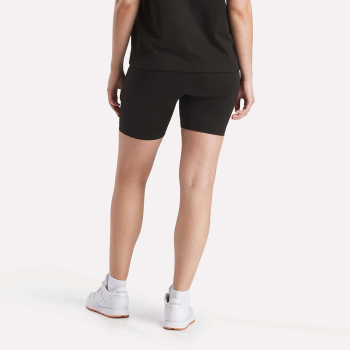 A person stands with their back to the camera, wearing a black t-shirt, white sneakers, and Reebok Women's Small Logo Cycle Shorts, fitted with a stretchy feel, against a plain light background.