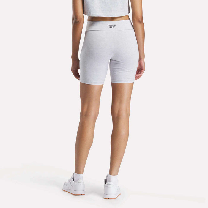 A woman in a gray crop top, Reebok Women's Small Logo Cycle Shorts in light gray, and white Reebok sneakers is photographed from behind against a plain white background.