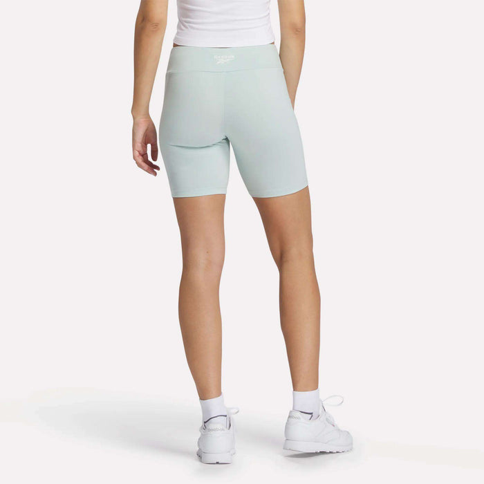 A person stands facing away, wearing Reebok Women's Small Logo Cycle Shorts in light blue, paired with a white top, white socks, and white sneakers against a plain white background.