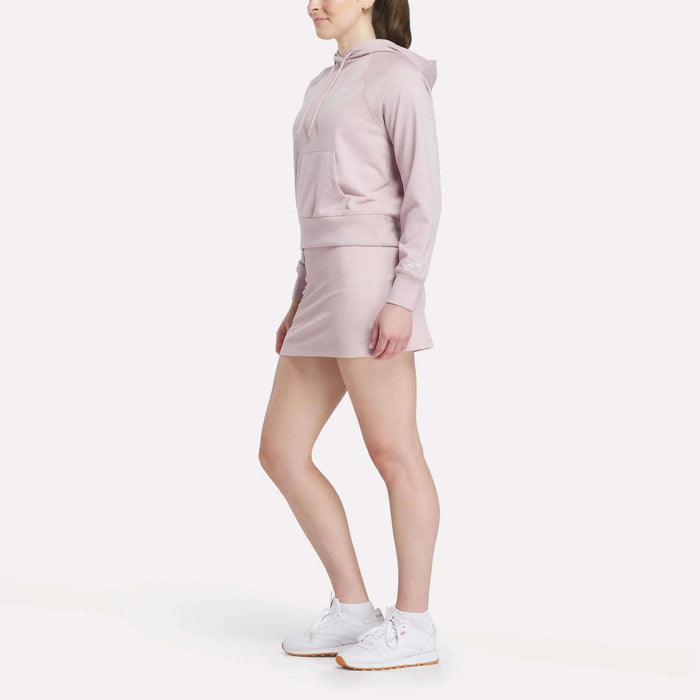 A woman models the Reebok Women's Small Vector Cropped Hoodie in light pink, paired with a matching skirt and white sneakers. She stands against a plain white background, facing slightly to the side with her hands at her sides.