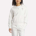 A woman wears the Reebok Women's Small Vector Full Zip Sweatshirt in white, paired with matching sweatpants, standing against a plain light background with one hand in her pocket and a relaxed expression.