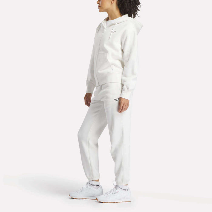 A woman wears the Reebok Women's Small Vector Full Zip Sweatshirt, paired with white sweatpants and sneakers. She stands against a plain white background, looking to the side with her hands relaxed by her sides.