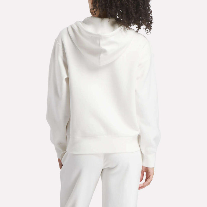 A person with curly hair, seen from behind, wears a Reebok Women's Small Vector Full Zip Sweatshirt in white and matching white pants. The hoodie is down, set against a plain light-colored background.