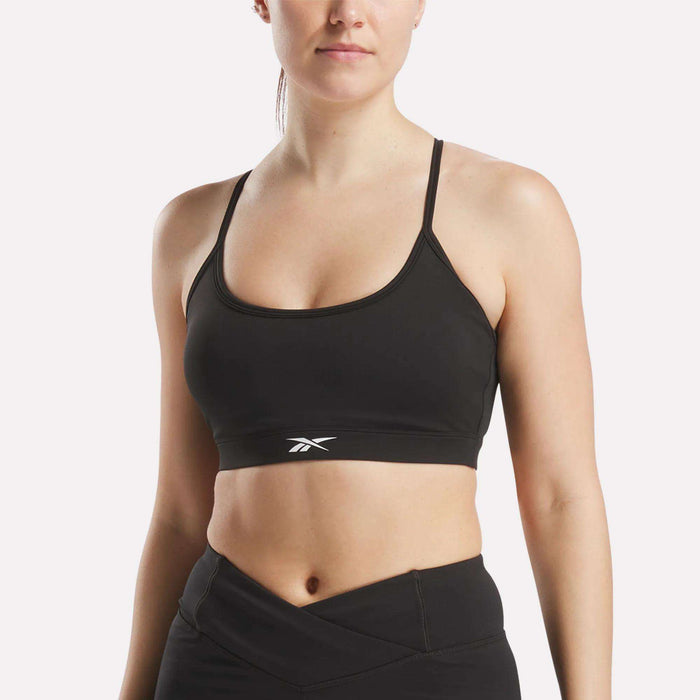 A woman wears the Reebok Women's Core Strappy Bra - Medium Support in black, paired with high-waisted athletic leggings, standing against a plain light background.