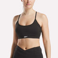 A woman wears the Reebok Women's Core Strappy Bra - Medium Support in black, paired with high-waisted athletic leggings, standing against a plain light background.