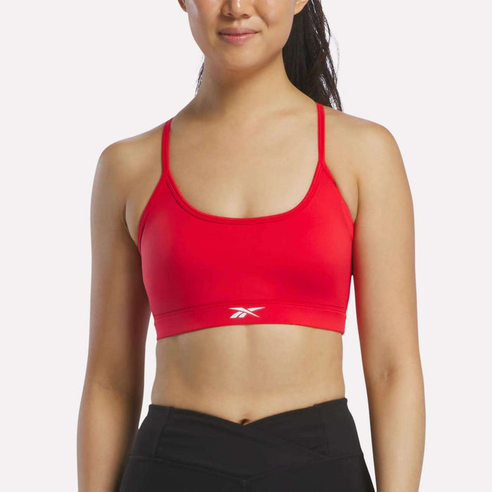 A woman in a Vector Red Reebok Women's Strappy Sports Bra and black athletic pants stands against a plain white background, smiling softly. Ideal for medium-impact workouts, the quick-drying bra features thin straps and a small white Reebok logo at the center.