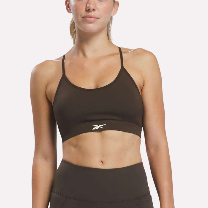A woman wearing the Reebok Women’s Strappy Sports Bra in black, paired with matching leggings, stands against a plain white background. Designed for medium-impact workouts, the sports bra features thin straps, quick-drying fabric, and a subtle Reebok logo at the center.