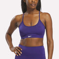 A woman stands with one hand on her hip against a plain white background, wearing a Reebok Women's Strappy Sports Bra in Bold Purple, designed to dry quickly and ideal for medium-impact workouts, paired with matching leggings.