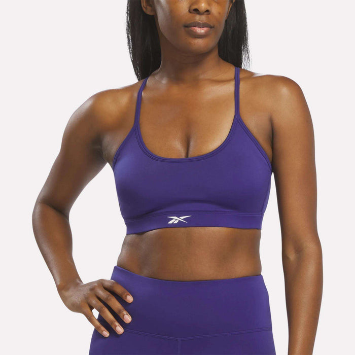A woman stands with one hand on her hip against a plain white background, wearing a Reebok Women's Strappy Sports Bra in Bold Purple, designed to dry quickly and ideal for medium-impact workouts, paired with matching leggings.