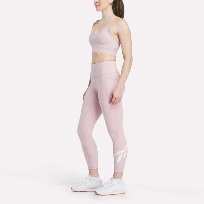 A woman models the Reebok Women's Back Bra and matching high-waisted leggings in light pink with white athletic shoes, standing sideways against a plain white background to highlight medium-impact support.