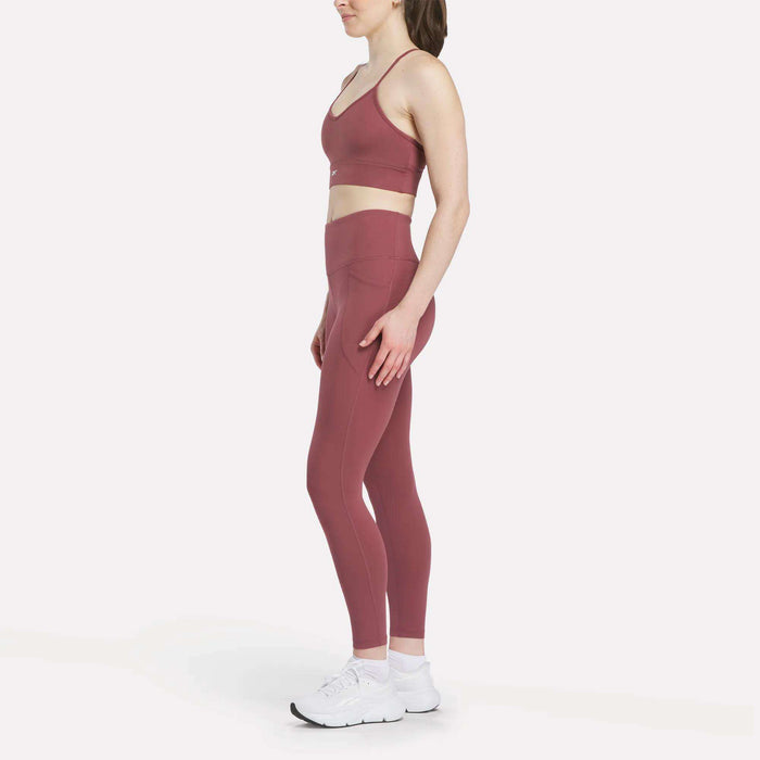 A woman stands in profile wearing a matching mauve Reebok Women's Back Bra and high-waisted leggings, paired with white sneakers, against a plain white background.