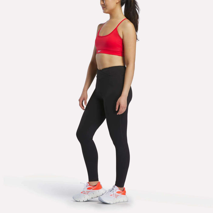 A woman stands sideways against a plain white background, wearing the Reebok Women's Strappy Sports Bra in red—ideal for medium-impact workouts—paired with black leggings and white athletic shoes featuring orange accents.