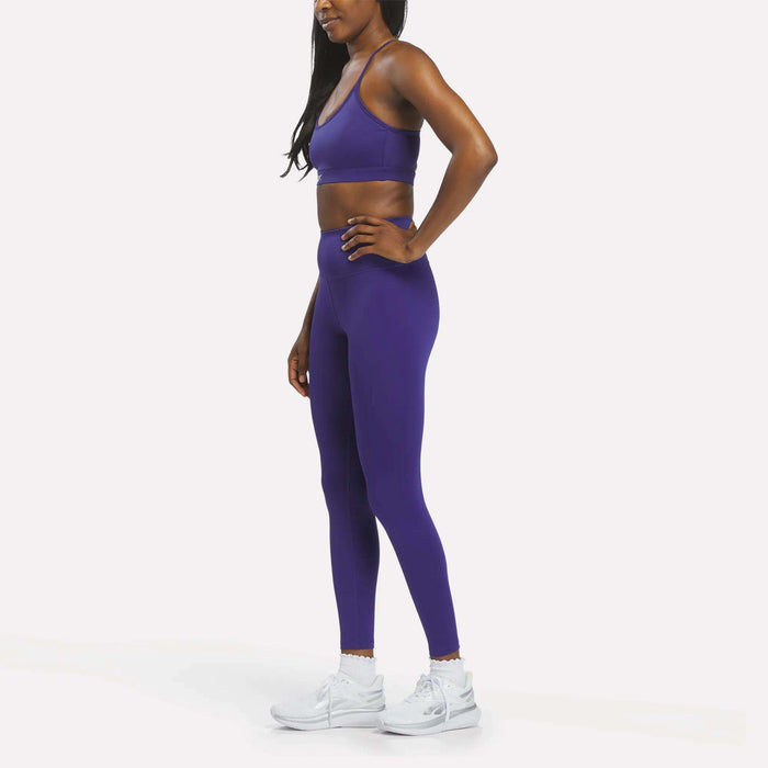 A woman stands in profile, wearing the Reebok Women's Strappy Sports Bra in purple paired with matching leggings and white athletic shoes against a plain white background—the quick-drying fabric of the sports bra makes it ideal for medium-impact workouts.
