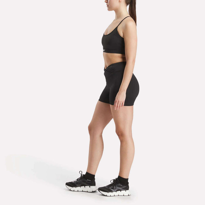 A woman stands in side profile against a white background, wearing the Reebok Women's Core Strappy Bra - Medium Support—ideal for medium-impact workouts with quick-drying comfort.