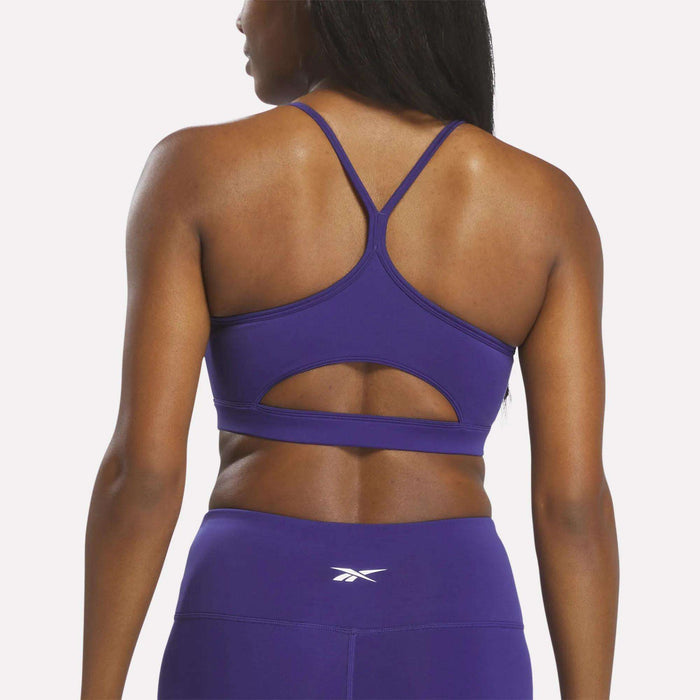 A woman is shown from the back against a plain white background, wearing the Reebok Women's Strappy Sports Bra in purple, featuring cutout detailing for medium-impact workouts and quick-drying comfort. She has paired it with matching high-waisted leggings.