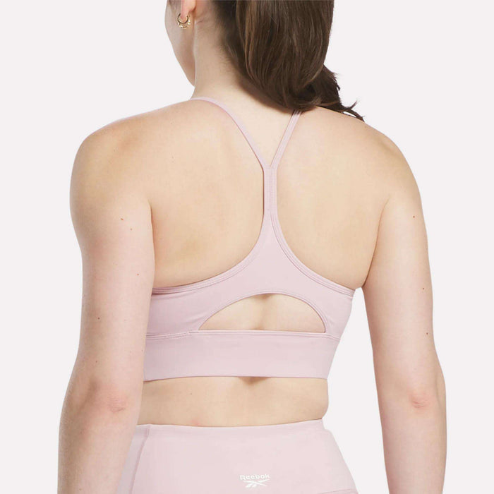 A woman with brown hair in a ponytail is seen from behind wearing the Reebok Women's Back Bra, which has thin straps, a keyhole cutout, and medium-impact support, paired with matching light pink leggings.
