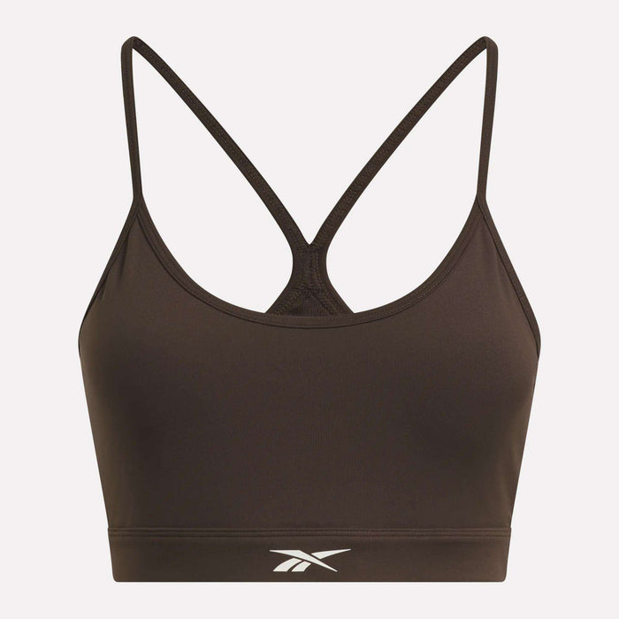 The Women’s Strappy Sports Bra by Reebok features a brown colorway with thin, crisscrossed straps in the back and a white Reebok logo centered on the front band. Designed for medium-impact workouts, this quick-drying sports bra is highlighted beautifully against a light pink background.