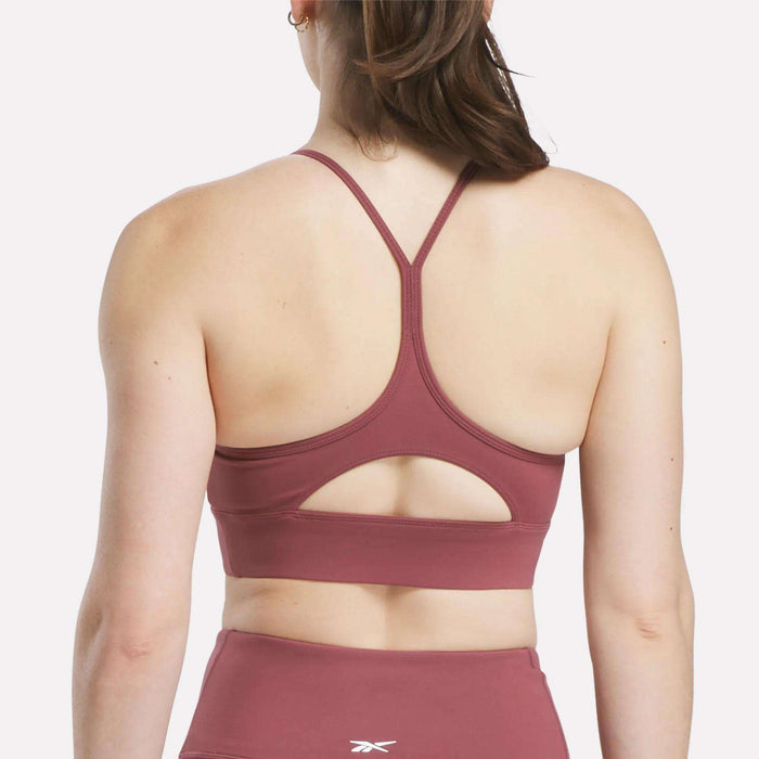 A woman is seen from behind wearing the Reebok Women's Back Bra in mauve with thin racerback straps and a small keyhole cutout, paired with matching high-waisted leggings.