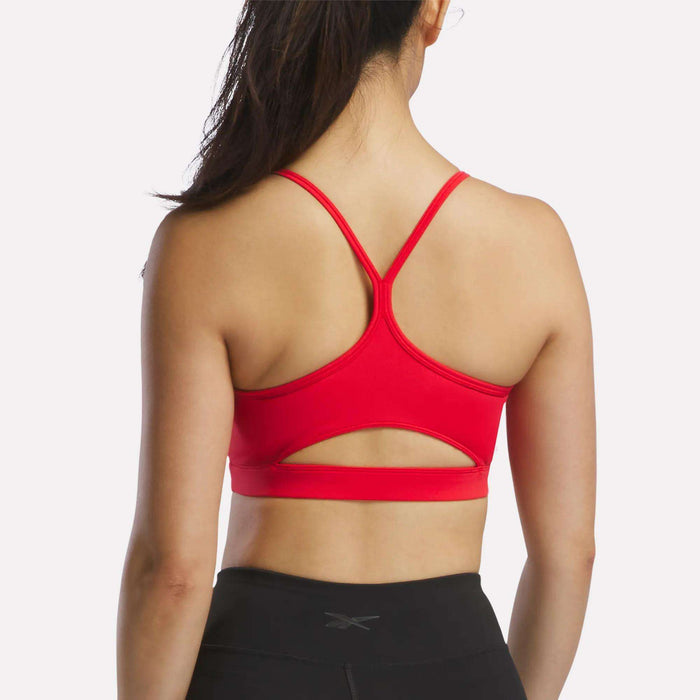 A woman is shown from the back wearing the Reebok Women's Strappy Sports Bra in red, featuring a stylish cutout design suitable for medium-impact workouts, paired with black athletic leggings. Her hair is tied in a ponytail, and she stands against a plain, light-colored background.