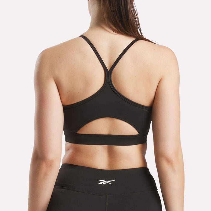 A woman is seen from the back wearing the Reebok Women's Core Strappy Bra - Medium Support, featuring thin, crisscrossed straps and a cutout detail, paired with black leggings with a small white Reebok logo on the waistband.