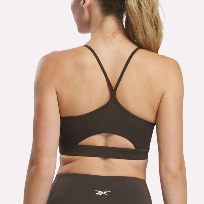A woman with blonde hair in a ponytail is shown from the back, wearing the Reebok Women’s Strappy Sports Bra featuring thin straps and a cut-out design. Perfect for medium-impact workouts, this bra is made from quick-drying fabric and is paired with matching athletic bottoms.