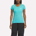 A woman in a fitted, short-sleeve Reebok Women's T-Shirt in Thati Blue and black pants stands facing forward against a plain white background.