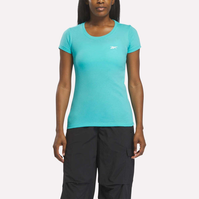 A woman in a fitted, short-sleeve Reebok Women's T-Shirt in Thati Blue and black pants stands facing forward against a plain white background.