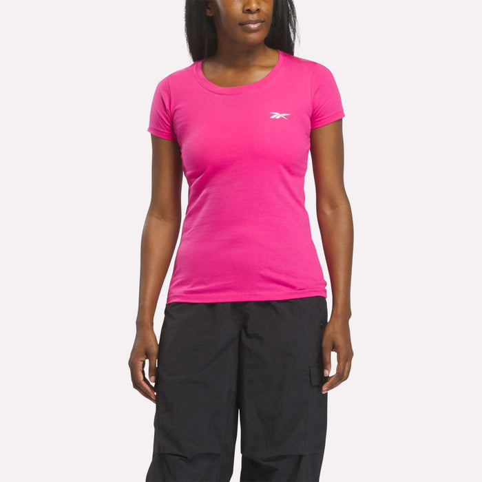 A woman stands against a white background wearing a raspberry Reebok women's t-shirt with black pants, her arms relaxed at her sides.
