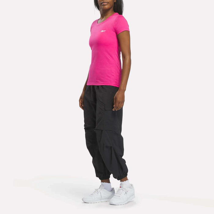A person stands against a white background wearing a bright pink Reebok Women's T-Shirt, black joggers, and white sneakers. Their hands are by their sides and their head is slightly turned.
