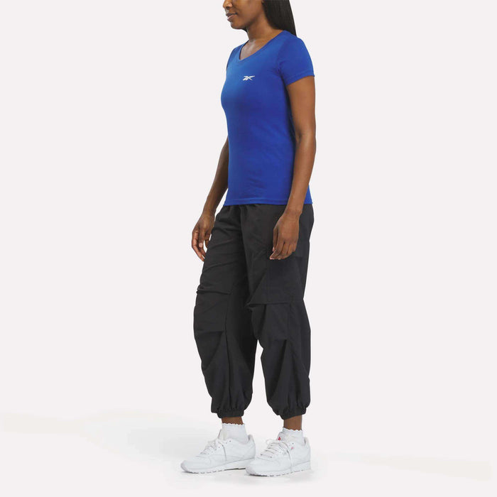 A woman faces left against a plain white background, wearing a fitted blue Reebok Women's T-Shirt, black cargo jogger pants, and white athletic sneakers.