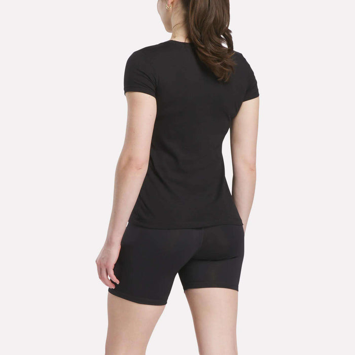 A person with long brown hair in a ponytail is standing and facing away, wearing a Reebok Women’s T-Shirt and black fitted shorts against a plain light background.