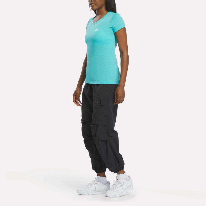 A person stands slightly sideways against a plain white background, wearing a fitted turquoise Reebok Women's T-Shirt and black cargo pants paired with white Reebok athletic sneakers.