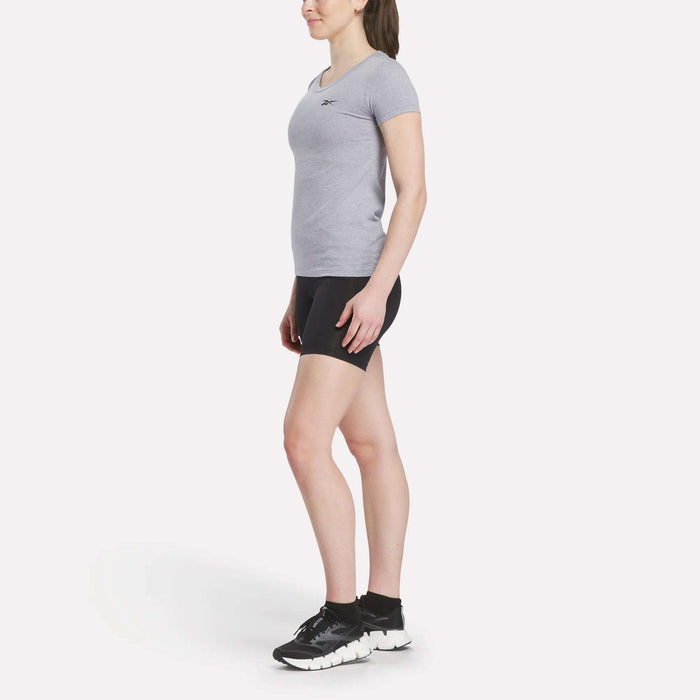 A woman poses in profile against a white background, wearing a fitted gray Women's Reebok T-Shirt, black shorts, black socks, and black athletic shoes.