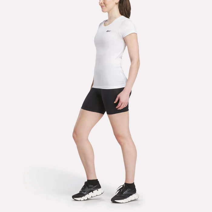 A woman stands against a plain background wearing a fitted Women's Reebok T-Shirt, black athletic shorts, black ankle socks, and black athletic shoes.