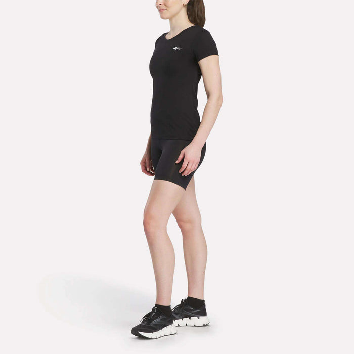 A woman poses in a fitted black Women's Reebok T-Shirt and black shorts with Reebok sneakers, set against a plain white background.