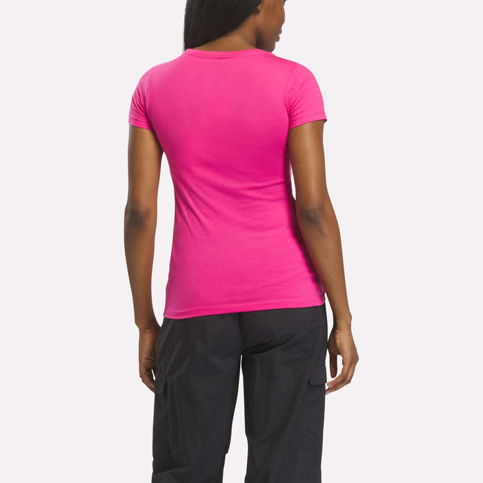 A woman faces away, wearing a Reebok Women's T-Shirt in bright pink with black pants, set against a plain white background.