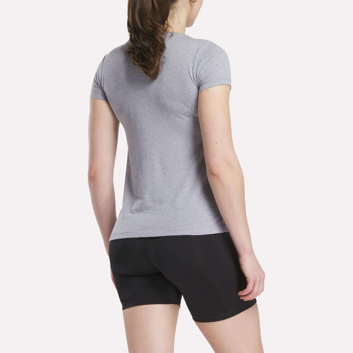 A woman with brown hair in a ponytail faces away from the camera, wearing a fitted Women's Reebok T-Shirt by Reebok and black shorts against a plain light background.