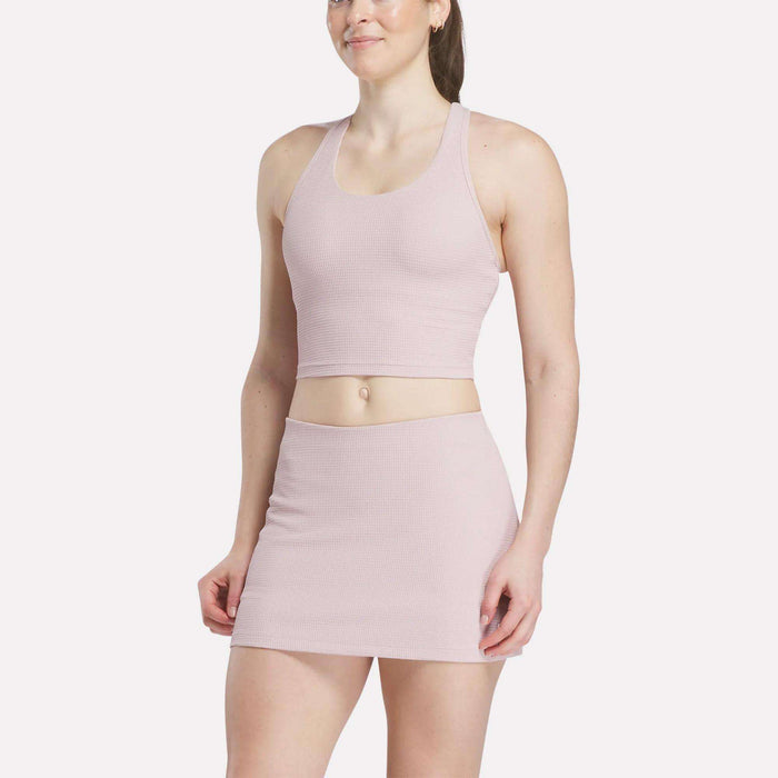 A woman in a light pink Women's Time Out Cropped Tank Top by Reebok and a matching high-waisted mini skirt stands against a plain white background, smiling slightly.