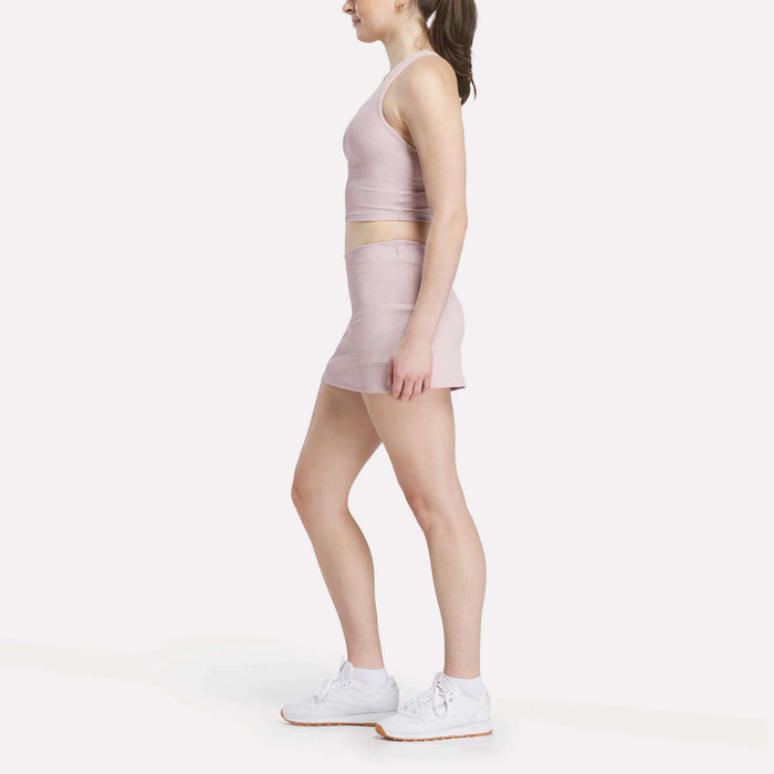 A woman stands in profile wearing a Reebok Women's Time Out Cropped Tank Top in light pink, a matching pink mini skirt, and white sneakers against a plain white background.