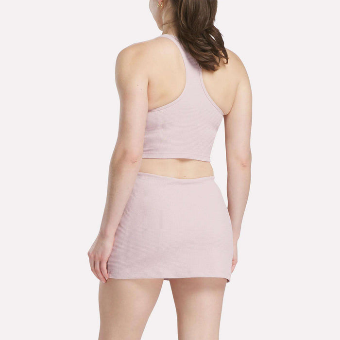 A woman with her hair tied back faces away, wearing the Reebok Women's Time Out Cropped Tank Top in light pink and a matching mini skirt set against a plain white background.