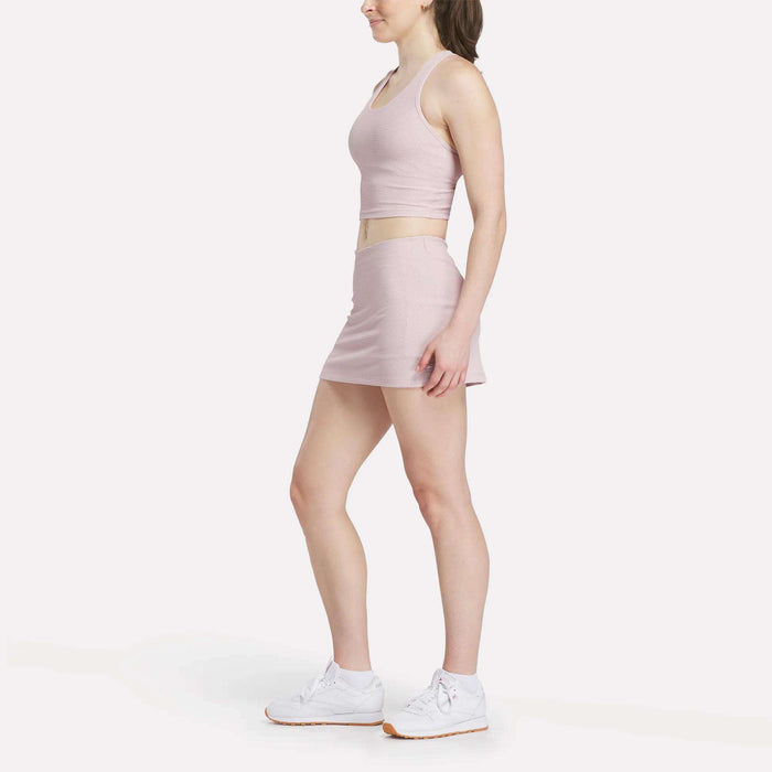 A woman in profile models a light pink sleeveless crop top with the women's Reebok Time Out Slim Tennis Skort and white Reebok sneakers, set against a plain white background.