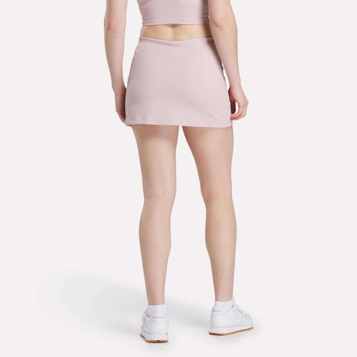 A woman stands with her back to the camera in a light pink Reebok Women's Time Out Slim Tennis Skort and matching crop top, styled with white sneakers, against a plain white background for a chic activewear look.