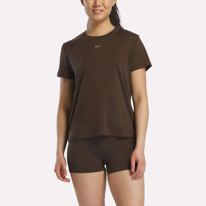A woman smiles against a white background, wearing an espresso Reebok Women's Training T-Shirt featuring a small logo, her hair pulled back and ready for her workout.