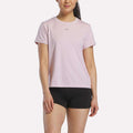 A woman wearing a Reebok Women's Training T-Shirt in Elemental Pink Melange and black shorts stands against a plain white background.
