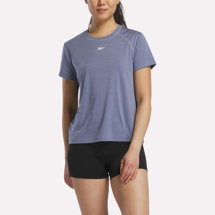 A woman wearing a hazy blue Reebok Women's Training T-Shirt and black shorts stands against a plain white background.