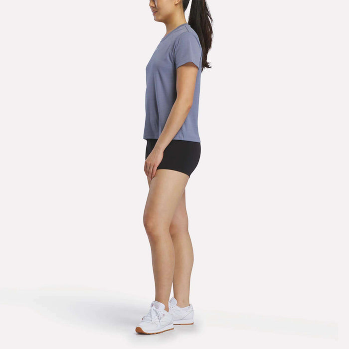 A woman is shown in profile, wearing a gray Reebok Women's Training T-Shirt, black shorts, and white sneakers against a plain white background.